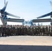 A large group of military personnel, predominantly in Marine Corps MARPAT camouflage uniforms with a smaller number in flight suits, pose for a formal group photograph on a flight line tarmac. They are standing in front of a V-22 Osprey tiltrotor aircraft with its rotors in the horizontal (airplane mode) position. The setting appears to be a military air station on a clear, sunny day.