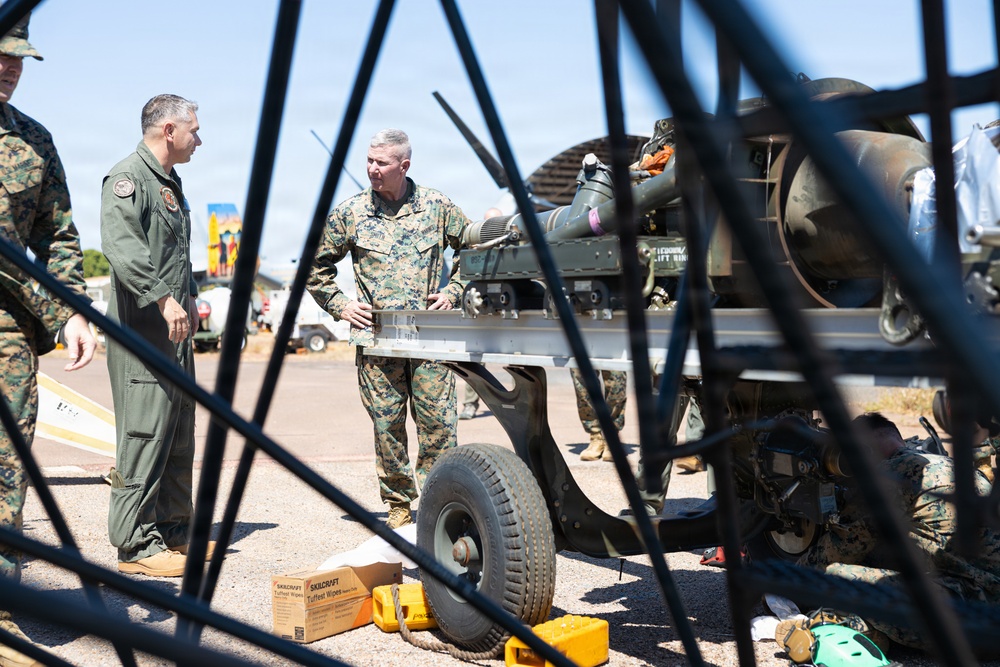 The image shows U.S. Marine Corps personnel in a maintenance or inspection setting, likely on a military flight line or airfield. Two senior personnel — one in a flight suit and one in MARPAT camouflage utilities — appear to be receiving a briefing or conducting an inspection of military equipment on a wheeled cart or trailer. The scene is partially obscured by dark structural elements in the foreground, suggesting the photo was taken through or around aircraft components.