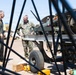 The image shows U.S. Marine Corps personnel in a maintenance or inspection setting, likely on a military flight line or airfield. Two senior personnel — one in a flight suit and one in MARPAT camouflage utilities — appear to be receiving a briefing or conducting an inspection of military equipment on a wheeled cart or trailer. The scene is partially obscured by dark structural elements in the foreground, suggesting the photo was taken through or around aircraft components.