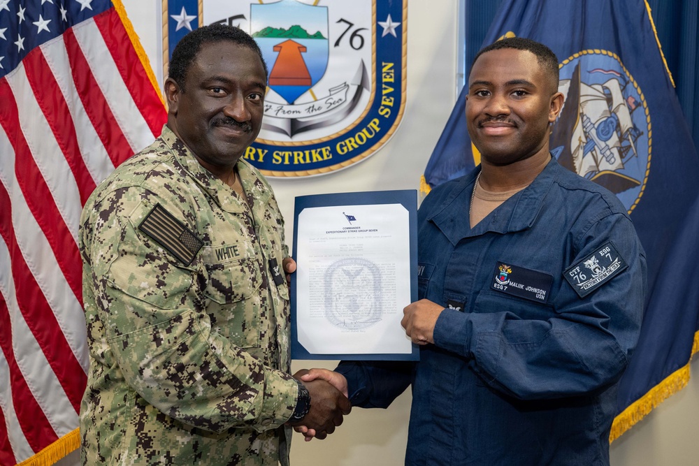 Two U.S. Navy personnel are posing for a formal recognition photo, shaking hands while jointly holding an official certificate or commendation letter. The setting is an official military backdrop featuring the Expeditionary Strike Group Seven seal, a U.S. flag, and a Navy unit flag. The occasion appears to be a formal award or recognition ceremony.
