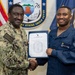Two U.S. Navy personnel are posing for a formal recognition photo, shaking hands while jointly holding an official certificate or commendation letter. The setting is an official military backdrop featuring the Expeditionary Strike Group Seven seal, a U.S. flag, and a Navy unit flag. The occasion appears to be a formal award or recognition ceremony.