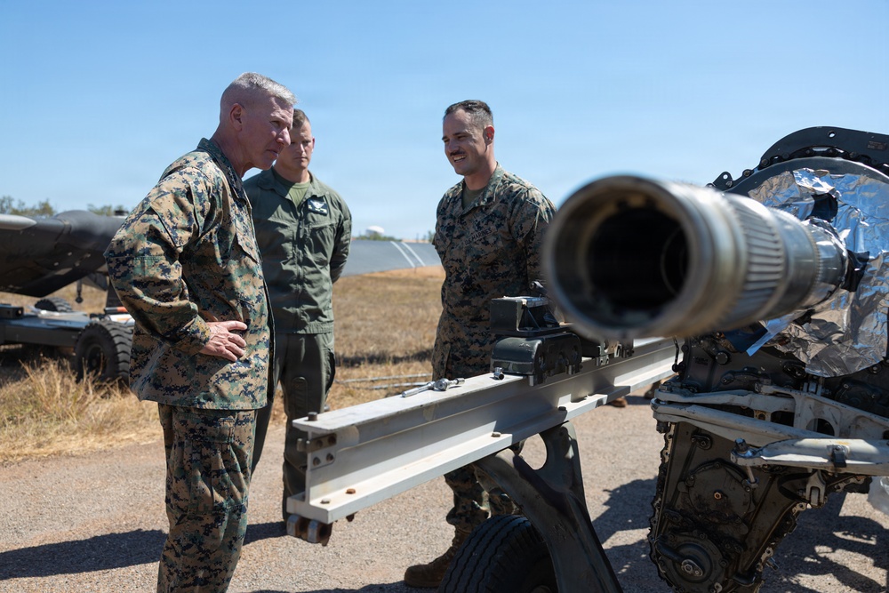 Three military personnel are gathered around what appears to be a wheeled artillery piece or weapon system in an outdoor setting. Two individuals are wearing USMC digital camouflage (MARPAT) uniforms, while one is wearing an olive/sage green flight suit. They appear to be engaged in a briefing or equipment inspection.