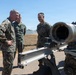 Three military personnel are gathered around what appears to be a wheeled artillery piece or weapon system in an outdoor setting. Two individuals are wearing USMC digital camouflage (MARPAT) uniforms, while one is wearing an olive/sage green flight suit. They appear to be engaged in a briefing or equipment inspection.