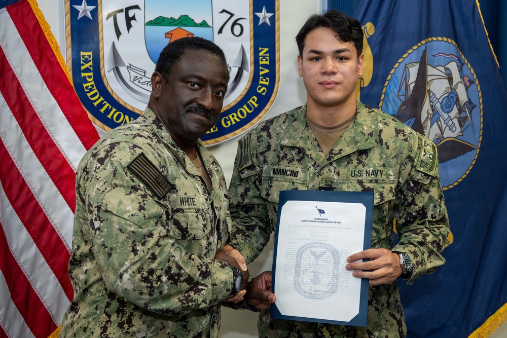 Two U.S. Navy personnel in NWU Type III (digital camouflage) uniforms pose for a formal photo. The sailor on the left, identified by name tape as 'WHITE,' is presenting a certificate or commendation document to the sailor on the right, identified as 'MANCINI.' The document header reads 'Commander Expeditionary Strike Group Seven,' and both are shaking hands in a ceremonial manner.