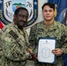 Two U.S. Navy personnel in NWU Type III (digital camouflage) uniforms pose for a formal photo. The sailor on the left, identified by name tape as 'WHITE,' is presenting a certificate or commendation document to the sailor on the right, identified as 'MANCINI.' The document header reads 'Commander Expeditionary Strike Group Seven,' and both are shaking hands in a ceremonial manner.