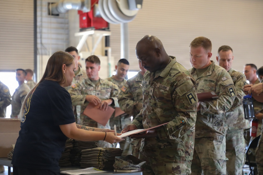 A group of U.S. Army soldiers in OCP (Operational Camouflage Pattern) uniforms are lined up at a processing or in-processing station inside what appears to be a military hangar or warehouse facility. A civilian woman in a navy blue t-shirt is assisting soldiers at a table, reviewing documents with them. Folded clothing items are visible on the table, suggesting this may be a uniform issue or administrative processing event.