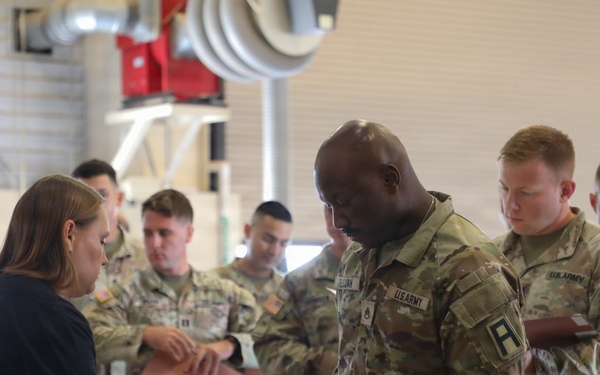 A group of U.S. Army soldiers in OCP (Operational Camouflage Pattern) uniforms are lined up at a processing or in-processing station inside what appears to be a military hangar or warehouse facility. A civilian woman in a navy blue t-shirt is assisting soldiers at a table, reviewing documents with them. Folded clothing items are visible on the table, suggesting this may be a uniform issue or administrative processing event.