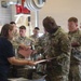 A group of U.S. Army soldiers in OCP (Operational Camouflage Pattern) uniforms are lined up at a processing or in-processing station inside what appears to be a military hangar or warehouse facility. A civilian woman in a navy blue t-shirt is assisting soldiers at a table, reviewing documents with them. Folded clothing items are visible on the table, suggesting this may be a uniform issue or administrative processing event.