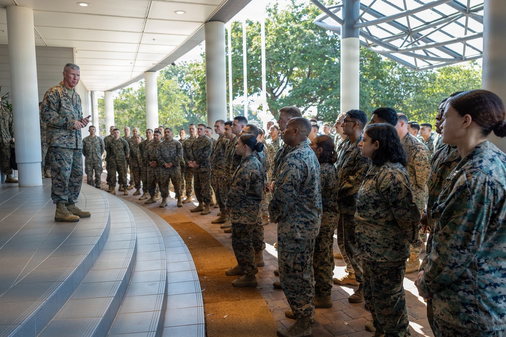 A senior military officer in U.S. Marine Corps digital camouflage (MARPAT) uniform is addressing a large formation of Marines under a covered outdoor walkway. The group of approximately 30-40 personnel, both male and female, stands in an attentive formation listening to the briefing. The setting appears to be a modern military installation or headquarters building.