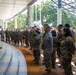 A senior military officer in U.S. Marine Corps digital camouflage (MARPAT) uniform is addressing a large formation of Marines under a covered outdoor walkway. The group of approximately 30-40 personnel, both male and female, stands in an attentive formation listening to the briefing. The setting appears to be a modern military installation or headquarters building.