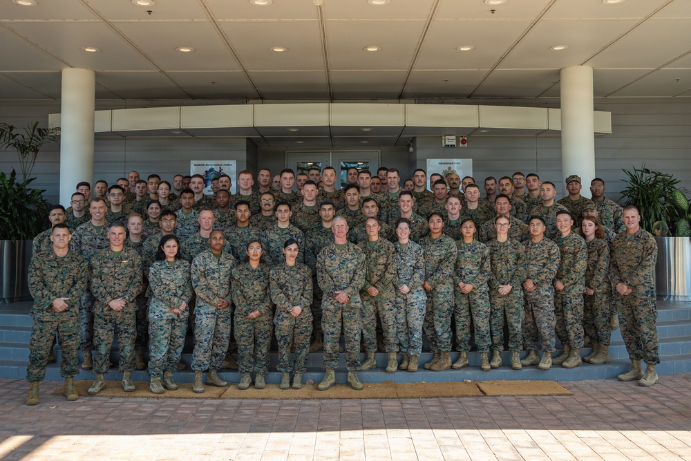A large group of U.S. Marine Corps personnel posed for a formal unit photograph in front of a modern military headquarters building. The group consists of both male and female Marines in woodland MARPAT camouflage uniforms, arranged in multiple rows on steps. Two signs visible in the background reference 'Marine Rotational Force' and 'Headquarters.'