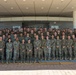 A large group of U.S. Marine Corps personnel posed for a formal unit photograph in front of a modern military headquarters building. The group consists of both male and female Marines in woodland MARPAT camouflage uniforms, arranged in multiple rows on steps. Two signs visible in the background reference 'Marine Rotational Force' and 'Headquarters.'