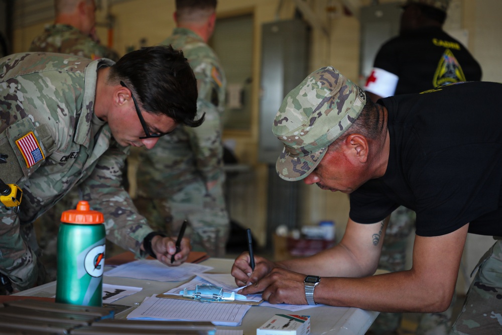 Two individuals in military attire are leaning over a table and writing on documents or forms, suggesting an administrative or processing activity. In the background, additional personnel in military uniforms are visible, including one individual wearing a red cross armband indicating a medical role. The setting appears to be an indoor military facility or staging area.