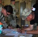 Two individuals in military attire are leaning over a table and writing on documents or forms, suggesting an administrative or processing activity. In the background, additional personnel in military uniforms are visible, including one individual wearing a red cross armband indicating a medical role. The setting appears to be an indoor military facility or staging area.