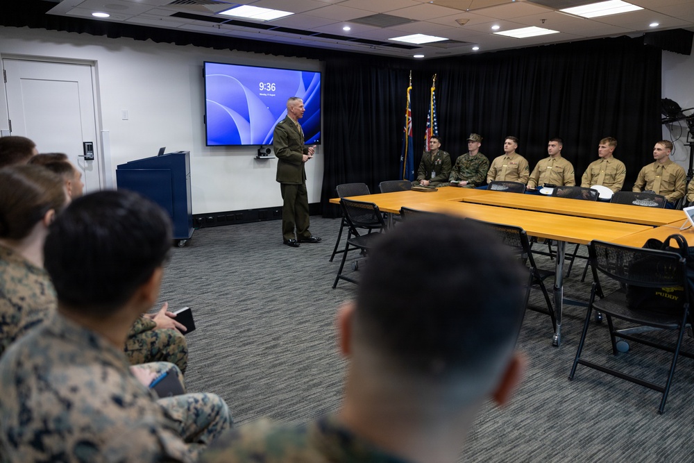 A military officer in olive green dress uniform is addressing a group of service members in a conference room setting. The briefing or meeting appears to be taking place at 9:36 AM, as shown on the display screen behind the speaker. Personnel are seated at tables and in chairs facing the standing officer.