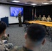 A military officer in olive green dress uniform is addressing a group of service members in a conference room setting. The briefing or meeting appears to be taking place at 9:36 AM, as shown on the display screen behind the speaker. Personnel are seated at tables and in chairs facing the standing officer.