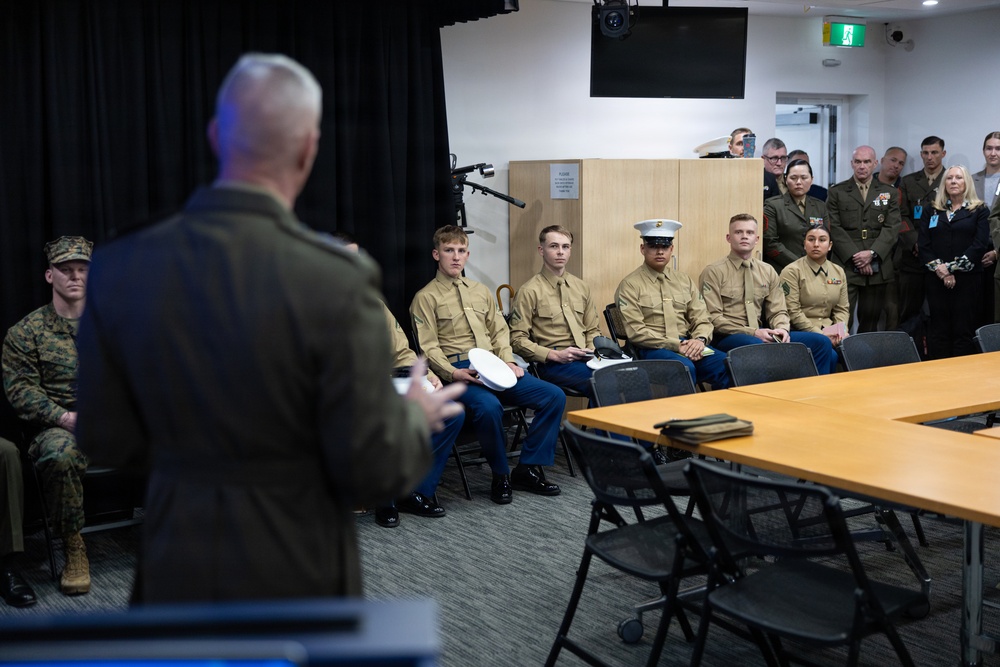A senior military officer, seen from behind, addresses a group of seated U.S. Marines in Service Charlie (tan shirt and blue trouser) uniforms in what appears to be a conference or briefing room. Additional personnel in combat utility uniforms and dress uniforms are visible along the sides and background. The setting suggests a formal military briefing, recognition ceremony, or official address.