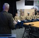 A senior military officer, seen from behind, addresses a group of seated U.S. Marines in Service Charlie (tan shirt and blue trouser) uniforms in what appears to be a conference or briefing room. Additional personnel in combat utility uniforms and dress uniforms are visible along the sides and background. The setting suggests a formal military briefing, recognition ceremony, or official address.