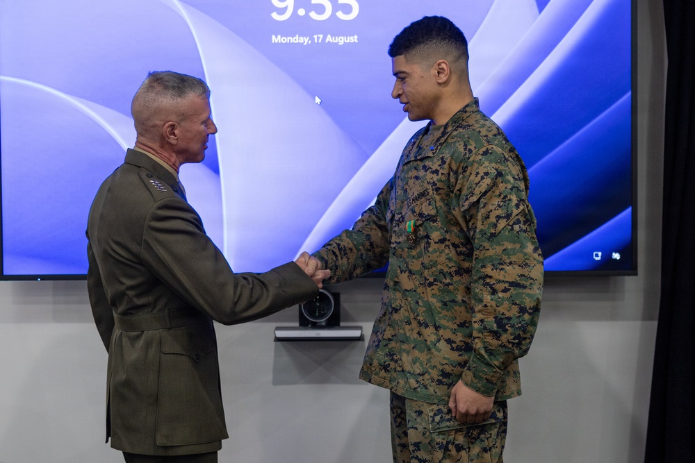 Two U.S. Marines are shaking hands in what appears to be a formal military ceremony, likely an award or recognition event. A senior officer in dress green uniform is presenting or congratulating a junior enlisted Marine in MARPAT camouflage utilities who has a medal pinned to his chest. The event takes place on Monday, 17 August at 9:55, as shown on the display screen behind them.