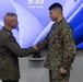 Two U.S. Marines are shaking hands in what appears to be a formal military ceremony, likely an award or recognition event. A senior officer in dress green uniform is presenting or congratulating a junior enlisted Marine in MARPAT camouflage utilities who has a medal pinned to his chest. The event takes place on Monday, 17 August at 9:55, as shown on the display screen behind them.