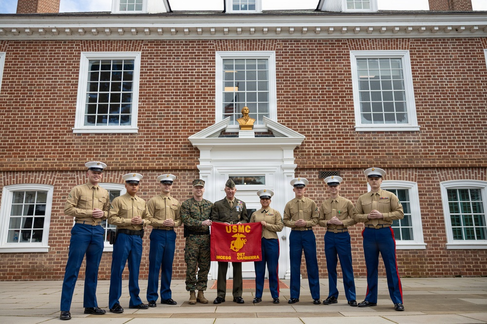 A group of nine United States Marine Corps personnel pose for a formal photograph in front of a red brick colonial-style building. Two central figures hold a red USMC guidon flag reading 'MCESO DET CANBERRA,' suggesting this is a detachment photo at or near the Marine Corps Embassy Security Group detachment in Canberra. Personnel are wearing a mix of Service Charlie uniforms (tan shirts with blue trousers) and dress uniforms.