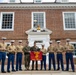 A group of nine United States Marine Corps personnel pose for a formal photograph in front of a red brick colonial-style building. Two central figures hold a red USMC guidon flag reading 'MCESO DET CANBERRA,' suggesting this is a detachment photo at or near the Marine Corps Embassy Security Group detachment in Canberra. Personnel are wearing a mix of Service Charlie uniforms (tan shirts with blue trousers) and dress uniforms.