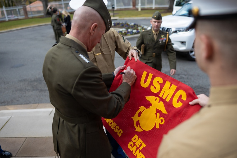 A senior U.S. Marine Corps officer is signing a red USMC banner or flag, which bears the Eagle, Globe, and Anchor emblem and text reading 'USMC' and 'DET CANN' (likely 'DET CANADA' or similar detachment designation). The scene appears to take place outdoors in a military installation setting, with several other uniformed Marines present. This appears to be a ceremonial or commemorative signing event.