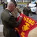A senior U.S. Marine Corps officer is signing a red USMC banner or flag, which bears the Eagle, Globe, and Anchor emblem and text reading 'USMC' and 'DET CANN' (likely 'DET CANADA' or similar detachment designation). The scene appears to take place outdoors in a military installation setting, with several other uniformed Marines present. This appears to be a ceremonial or commemorative signing event.