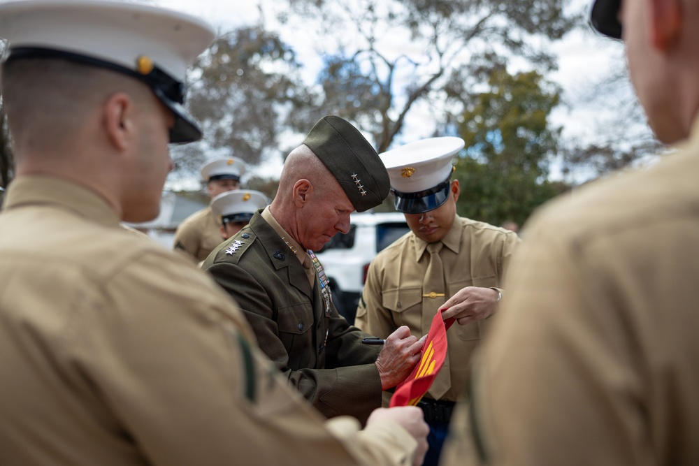 A senior military officer wearing a dark green service uniform with four-star rank insignia and multiple medals is signing a red and gold flag or banner. He is surrounded by several Marines in tan service uniforms and white dress covers. The setting appears to be an outdoor military event with trees visible in the background.