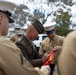 A senior military officer wearing a dark green service uniform with four-star rank insignia and multiple medals is signing a red and gold flag or banner. He is surrounded by several Marines in tan service uniforms and white dress covers. The setting appears to be an outdoor military event with trees visible in the background.