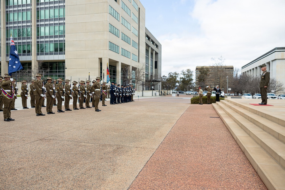 A formal military parade or honor guard ceremony is taking place outdoors at what appears to be a government or defense facility in Australia, identifiable by the Australian flag visible on the left. Two distinct formations of military personnel in different uniforms — army (olive/khaki) and air force (dark blue) — are lined up facing a senior officer standing on steps to the right. A red carpet leads from the steps toward the formations, suggesting a formal reception or inspection ceremony.