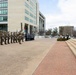 A formal military parade or honor guard ceremony is taking place outdoors at what appears to be a government or defense facility in Australia, identifiable by the Australian flag visible on the left. Two distinct formations of military personnel in different uniforms — army (olive/khaki) and air force (dark blue) — are lined up facing a senior officer standing on steps to the right. A red carpet leads from the steps toward the formations, suggesting a formal reception or inspection ceremony.