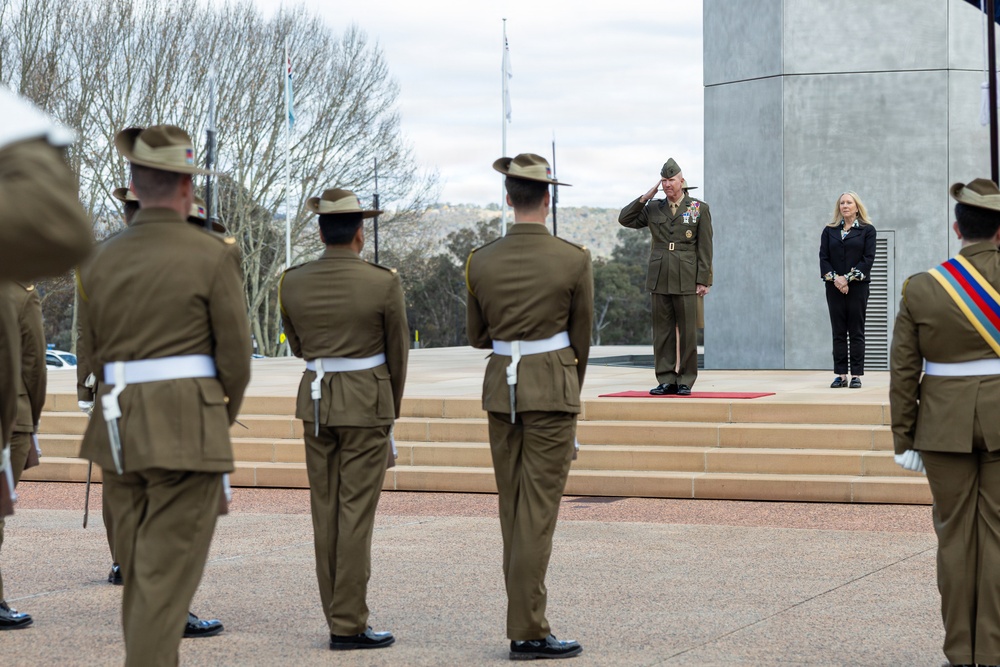 A formal military ceremony is taking place at what appears to be an outdoor monument or memorial, likely in Australia based on the distinctive Australian Army uniforms and slouch hats worn by the honor guard. A senior officer in a different uniform (appearing to be US Marine Corps based on the garrison cap and olive uniform) stands on a red carpet and renders a salute. A civilian woman in a dark suit stands nearby observing the proceedings.