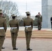 A formal military ceremony is taking place at what appears to be an outdoor monument or memorial, likely in Australia based on the distinctive Australian Army uniforms and slouch hats worn by the honor guard. A senior officer in a different uniform (appearing to be US Marine Corps based on the garrison cap and olive uniform) stands on a red carpet and renders a salute. A civilian woman in a dark suit stands nearby observing the proceedings.