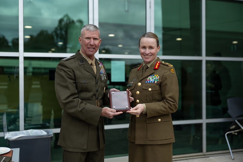Two senior military officers in dress uniforms are jointly holding a commemorative plaque or award box during what appears to be an official ceremony or exchange. The officer on the left wears a US Marine Corps uniform with extensive medal ribbons, while the officer on the right wears what appears to be an Australian Army uniform with rank insignia and medal ribbons. Both are smiling, suggesting a positive diplomatic or ceremonial occasion.