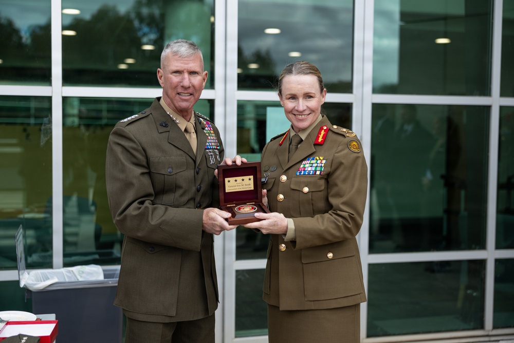 Two senior military officers, one appearing to be a U.S. Marine Corps general and the other an Australian Army officer, pose together while jointly holding a wooden presentation box containing a commemorative coin or medal. The occasion appears to be a formal military exchange or ceremony, likely involving a gift presentation between allied forces. Both officers are in dress uniforms displaying numerous medals and insignia indicating high rank.