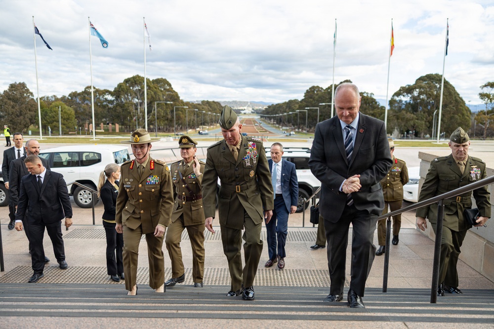 A formal military and diplomatic delegation is ascending steps at what appears to be the Australian War Memorial or a government building in Canberra, Australia. The group includes personnel in Australian Army uniforms, US Marine Corps uniforms, and civilian business attire. The backdrop shows the ANZAC Parade leading toward Parliament House, with multiple national flags visible.