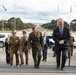 A formal military and diplomatic delegation is ascending steps at what appears to be the Australian War Memorial or a government building in Canberra, Australia. The group includes personnel in Australian Army uniforms, US Marine Corps uniforms, and civilian business attire. The backdrop shows the ANZAC Parade leading toward Parliament House, with multiple national flags visible.