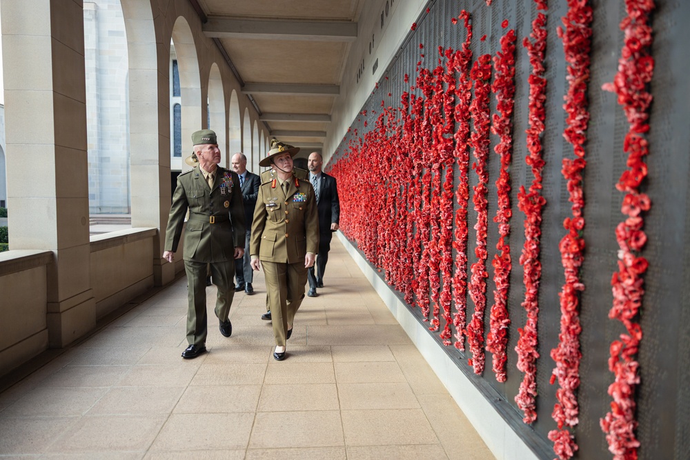 Two senior military officers in uniform walk alongside a large memorial wall densely covered with red poppies, accompanied by several civilians in the background. The setting appears to be a war memorial with a colonnaded sandstone corridor, consistent with the Australian War Memorial in Canberra. The red poppies on the wall serve as a tribute to fallen soldiers.