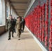 Two senior military officers in uniform walk alongside a large memorial wall densely covered with red poppies, accompanied by several civilians in the background. The setting appears to be a war memorial with a colonnaded sandstone corridor, consistent with the Australian War Memorial in Canberra. The red poppies on the wall serve as a tribute to fallen soldiers.