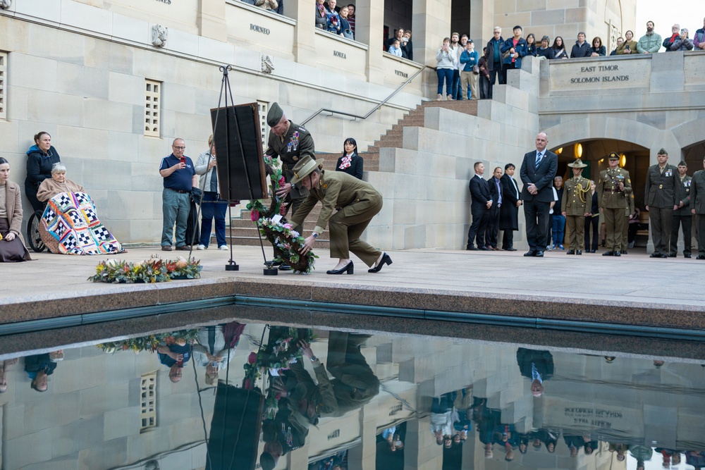 A military commemoration ceremony is taking place at what appears to be the Australian War Memorial in Canberra, featuring a wreath-laying ceremony beside a reflective pool. Two uniformed military personnel are placing a floral wreath near the pool, while crowds of onlookers observe from surrounding areas including elevated steps. The stone walls bear inscriptions of conflict names including 'Borneo', 'Korea', 'Vietnam', 'East Timor', and 'Solomon Islands'.