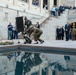 A military commemoration ceremony is taking place at what appears to be the Australian War Memorial in Canberra, featuring a wreath-laying ceremony beside a reflective pool. Two uniformed military personnel are placing a floral wreath near the pool, while crowds of onlookers observe from surrounding areas including elevated steps. The stone walls bear inscriptions of conflict names including 'Borneo', 'Korea', 'Vietnam', 'East Timor', and 'Solomon Islands'.