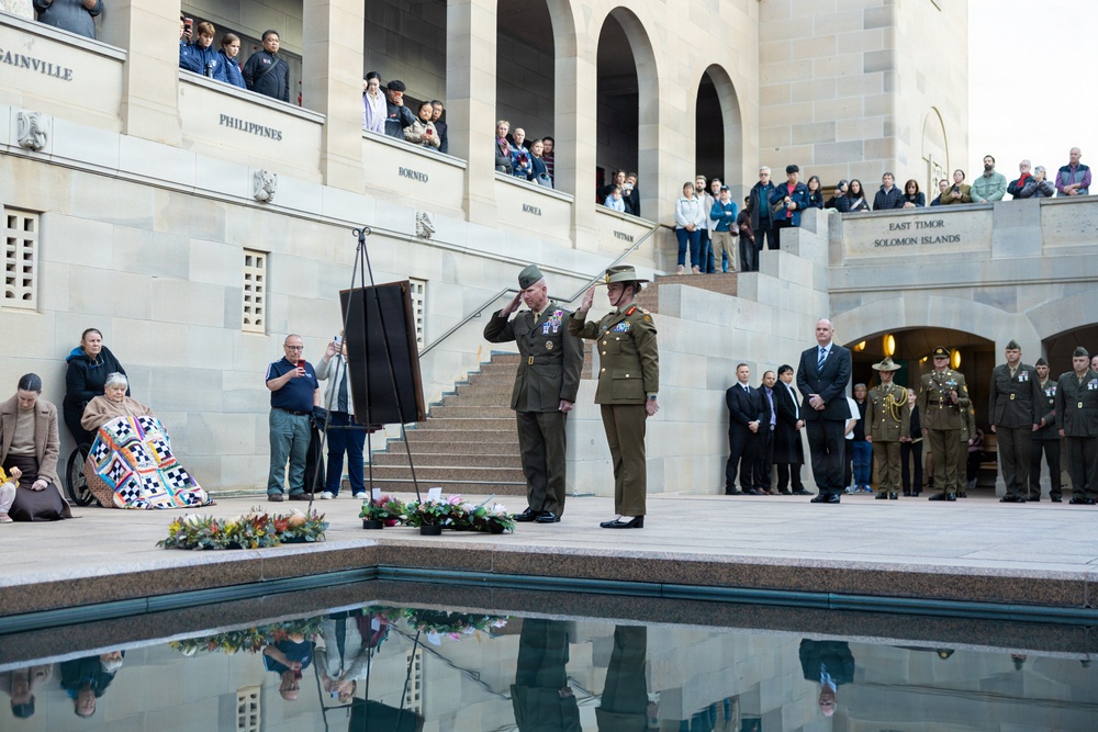 A formal military commemoration ceremony is taking place at what appears to be the Australian War Memorial in Canberra. Two senior military officers in olive green uniforms are saluting beside a lectern, with floral wreaths laid on the ground near a reflective pool. A large crowd of spectators, including civilians and uniformed personnel, observe from surrounding terraces and steps.