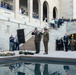 A formal military commemoration ceremony is taking place at what appears to be the Australian War Memorial in Canberra. Two senior military officers in olive green uniforms are saluting beside a lectern, with floral wreaths laid on the ground near a reflective pool. A large crowd of spectators, including civilians and uniformed personnel, observe from surrounding terraces and steps.