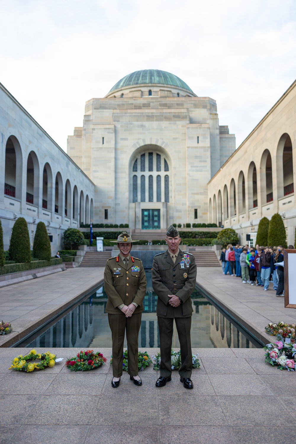 Two senior military officers — one in Australian Army uniform with a slouch hat, one in US Marine Corps dress uniform — stand at attention in front of the Pool of Reflection at the Australian War Memorial in Canberra. Floral wreaths are laid along the edge of the pool, indicating a commemorative ceremony has taken place. A group of visitors or students can be seen lined up along the right colonnade in the background.