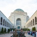 Two senior military officers — one in Australian Army uniform with a slouch hat, one in US Marine Corps dress uniform — stand at attention in front of the Pool of Reflection at the Australian War Memorial in Canberra. Floral wreaths are laid along the edge of the pool, indicating a commemorative ceremony has taken place. A group of visitors or students can be seen lined up along the right colonnade in the background.