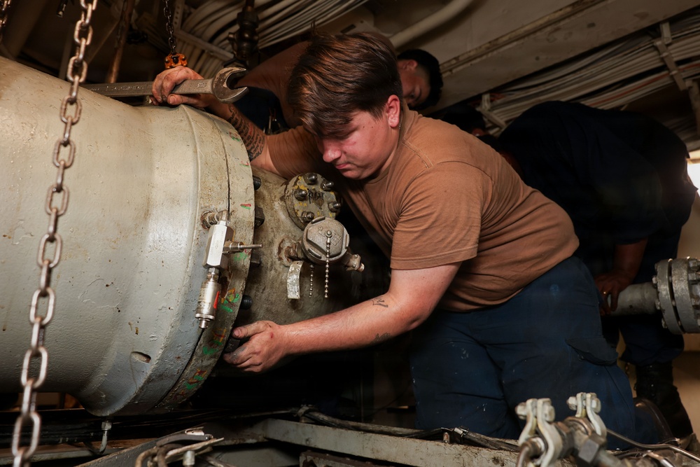 A person in a tan/brown military-style t-shirt is working on a large cylindrical piece of equipment, using a wrench and hands-on manipulation. The equipment appears to be a large ordnance or industrial cylinder secured by chains, with various fittings, valves, and connectors on its end cap. At least one other person is visible working in the background in a confined space.