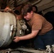 A person in a tan/brown military-style t-shirt is working on a large cylindrical piece of equipment, using a wrench and hands-on manipulation. The equipment appears to be a large ordnance or industrial cylinder secured by chains, with various fittings, valves, and connectors on its end cap. At least one other person is visible working in the background in a confined space.