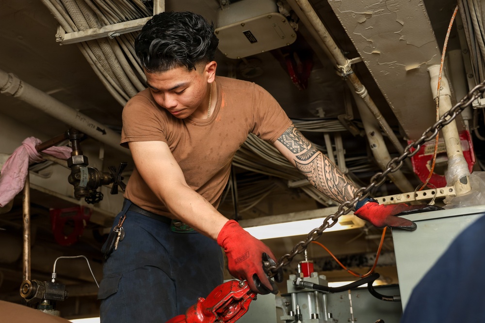 A young male technician or military sailor is working in a confined mechanical space, handling heavy chains and red-painted equipment. He is wearing red protective gloves, a brown t-shirt, and dark work pants, with a prominent tribal tattoo on his right forearm. The environment appears to be the interior of a naval vessel, characterized by pipes, cables, and industrial machinery.