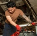 A young male technician or military sailor is working in a confined mechanical space, handling heavy chains and red-painted equipment. He is wearing red protective gloves, a brown t-shirt, and dark work pants, with a prominent tribal tattoo on his right forearm. The environment appears to be the interior of a naval vessel, characterized by pipes, cables, and industrial machinery.